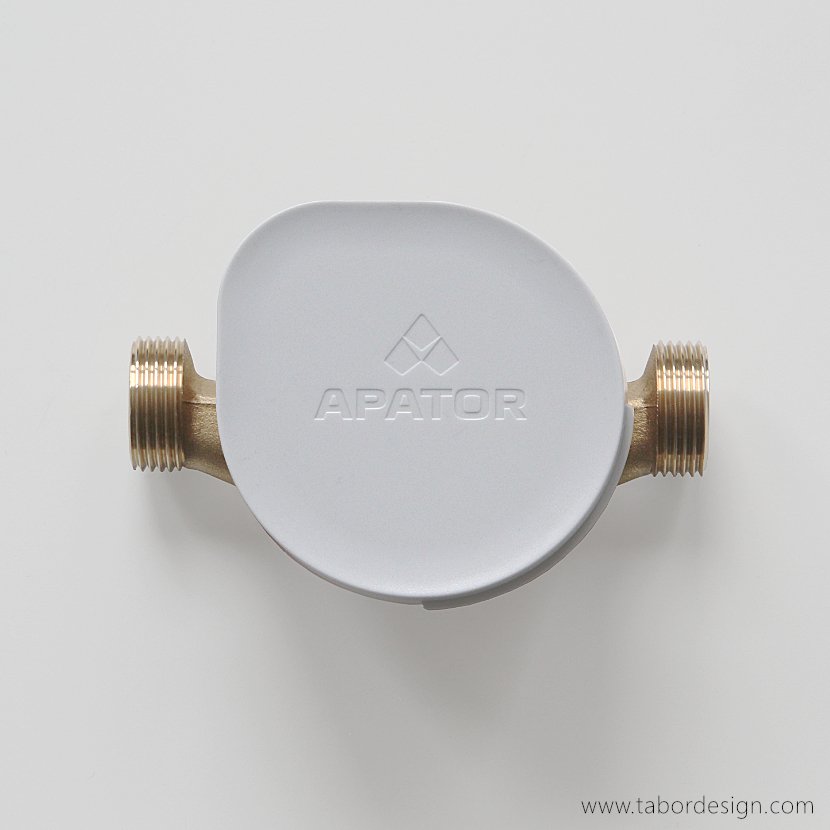 Wodomierz Apator widziany z góry. Tarcza liczydła zasłonięta klapką z logo firmy. Kształt klapki to kropla wody. Widoczne gwinty przyłączy.